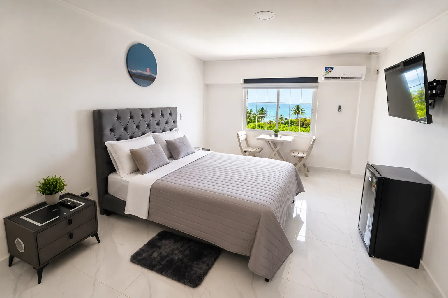 Habitación con cama doble de cabecero capitoneado y una mesa para dos junto a la ventana, con palmeras y el mar al fondo.